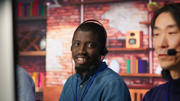 Portrait of smiling technical support specialist in call center office doing troubleshooting. Upbeat african american IT worker fixing customer connectivity issue, using headphones, camera A