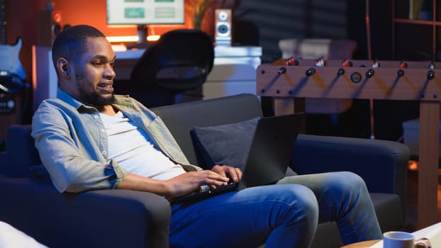 Adult guy typing on laptop laying on couch in stylish apartment, engaging in freelancing and online work. Black person multitasking efficiently and embracing flexibility of remote job. Camera B.