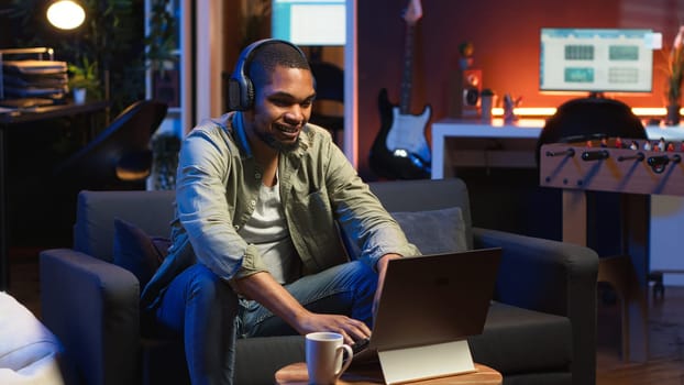 African American guy working on laptop celebrating achievement under neon lights, sitting on a couch in cozy apartment. Relaxed person listens to music, pleased with tasks success. Camera B.