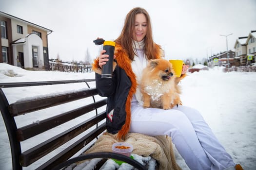 A woman sits on a bench, holding a drink and snacks while her fluffy Pomeranian rests beside her in the snow.