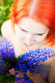 A woman with vibrant red hair gazes down at a bouquet of purple flowers. Surrounded by greenery, she enjoys the beauty of nature on a sunny day.