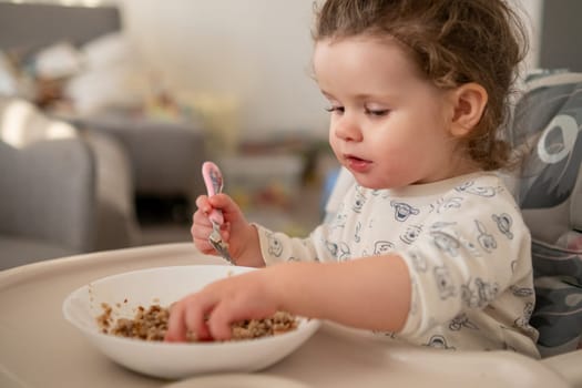 Little girl with curly hair focused on eating porridge in the kitchen. Messy and sweet moment of toddler mealtime.
