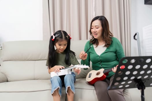 A mother guides her daughter in playing the ukulele, promoting music education and family connection in a warm home setting.
