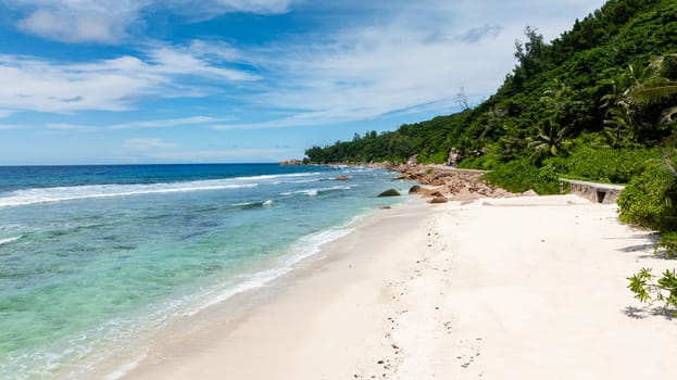 Shallow clear waters with patches of coral along a coastline bordered by a narrow white sandy beach. Seychelles, La Digue.