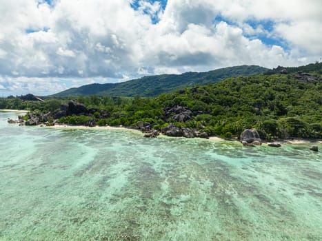 Aerial shot of lush green hills meeting the coast with cloudy skies overhead. La Digue, Seychelles.