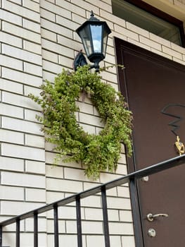 decorative wreath of fresh twigs, sage flowers and leaves with a purple ribbon hangs against the background of a light cypress wall next to the front door to the house. High quality photo