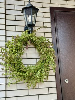 decorative wreath of fresh twigs, sage flowers and leaves with a purple ribbon hangs against the background of a light cypress wall next to the front door to the house. High quality photo