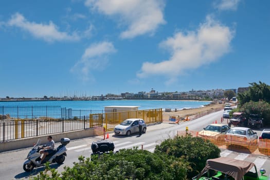 Rodas.Greece - june 05, 2025: A panoramic view of Rhodes bay, showing the beach, turquoise sea, and coastline with vehicles and some constructions. The blue sky with clouds adorns the scene.