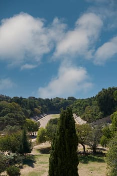 A panoramic view of the ancient stadium in Rhodes, Greece, with stands and lush greenery under a blue sky with clouds, ideal for heritage.