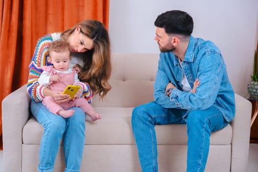 Tense family moment where a father looks upset while the mother shows cartoons to their baby on a smartphone, illustrating parenting conflict over screen time and digital content for children.