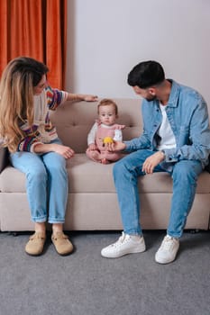 Vertical portrait Smiling parents enjoy playtime with their baby girl who holds toys while sitting on a sofa. A joyful family bonding moment filled with love, color, and warmth in a cozy home setting.