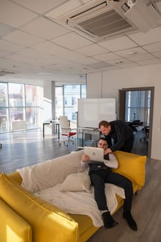 Two Caucasian men discussing work on a laptop while sitting on a sofa. Modern office