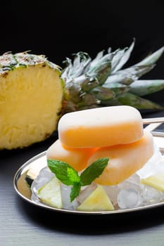 Pineapple sorbet ice cream popsicles on silver plate with ice cubes chopped pineapple slices, mint near cut in half pineapple on a black background. Pineapple sorbet ice cream popsicles. Vertical.