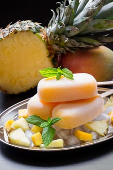 Exotic sorbet ice cream popsicles on silver plate with ice cubes chopped pineapple mango pieces, mint near half of pineapple, mango on a black background. Pineapple mango sorbet ice cream popsicles.
