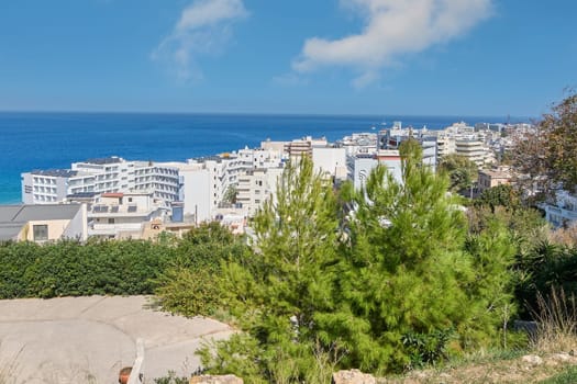 Rodas.Greece - june 07, 2025: Breathtaking elevated perspective of a bustling coastal Greek city, with numerous white structures nestled among lush greenery, extending towards the azure waters