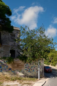 Rhodes. Greece - June 06, 2025: Urban scene capturing a vibrant mural decorating an old wall in Rhodes, with a lush tree in the foreground and a vehicle passing by. The combination of art and historic