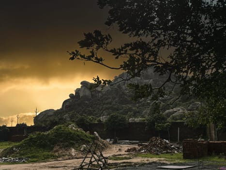 A dramatic landscape featuring rocky hills silhouetted against a vibrant, golden-hued sunset or sunrise sky. In the foreground, a semi-urban area is visible with sparse vegetation, a dirt path, and hints of construction materials, suggesting ongoing development. A tree branch frames the upper right, adding depth to the scene. This image evokes feelings of rugged nature, the beauty of twilight, and the juxtaposition of natural landscapes with human activity. Ideal for themes of nature, landscapes, sunsets, travel, and rural development. Dramatic Sunset Over Rocky Hills and Rural Landscape, Naga Hills, Kanke to Patratu Road, Ranchi, Jharkhand. High quality photo