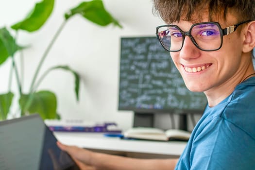 Cute teenage student in glasses sitting at a computer studying online, looking at the camera and smiling contentedly. High quality photo