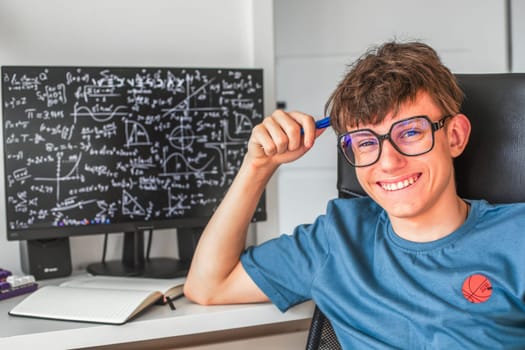 Cute teenage student in glasses sitting at a computer studying online, looking at the camera and smiling contentedly. High quality photo