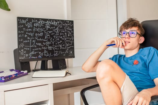 A teenage boy in glasses ponders solving math problems while sitting at a computer monitor. High quality photo