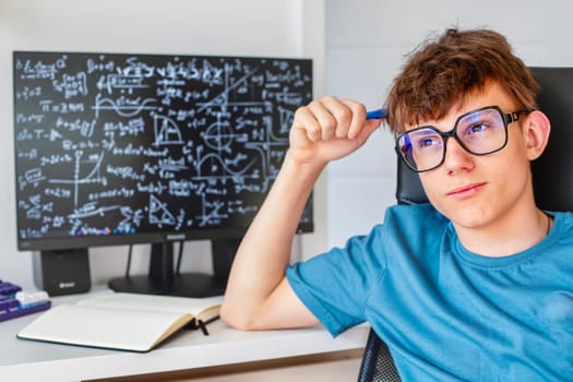 A teenage boy in glasses ponders solving math problems while sitting at a computer monitor. High quality photo