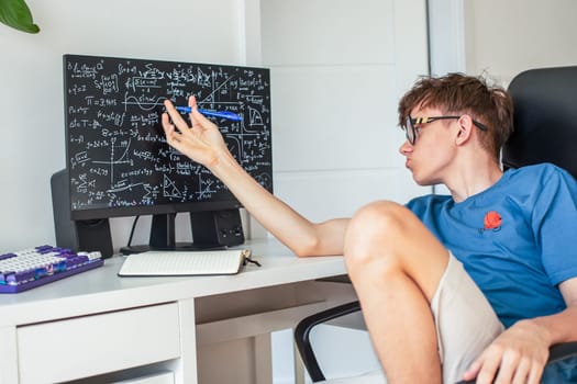 A teenage boy in glasses ponders solving math problems while sitting at a computer monitor. High quality photo