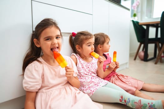 Three little girls sit on a floor, savoring bright ice pops.