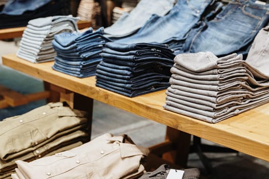 Three stacks of beautifully folded different colors new jeans lie diagonally on a wooden shelf table in a men's store,side view close-up.Concept time offline shopping.