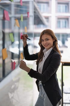 An asian businesswoman uses sticky notes on glass in a modern office space..