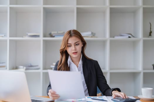 A young businesswoman carefully reviews documents while using calculator at desk..