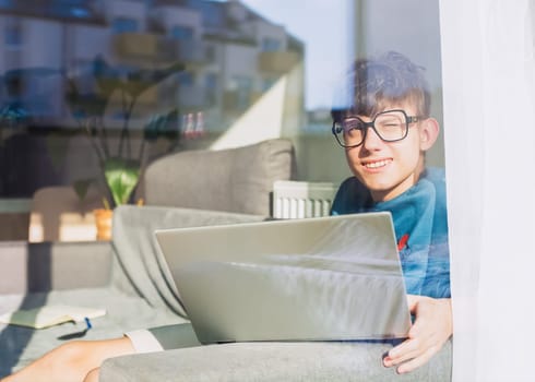 Happy student in glasses sitting with laptop and looking blinded by the sun through the window, student studying online. High quality photo