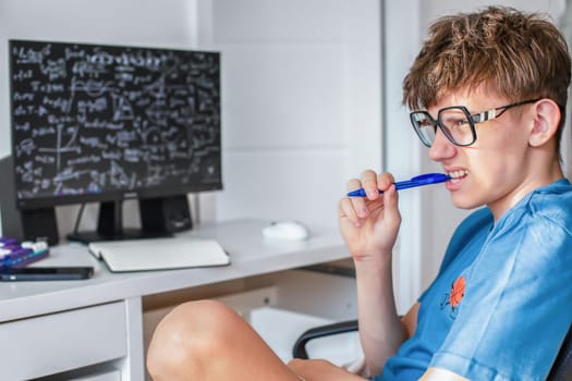 A student in glasses sits studying at a computer monitor with a pen in his teeth, and is tensely solving math problems. High quality photo