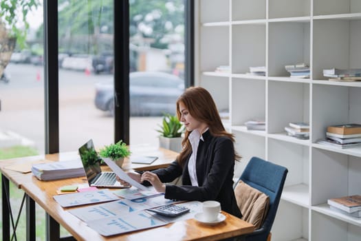 Young businesswoman reviews paperwork while working on laptop in office space.