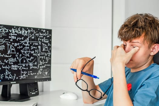 Tired and exhausted teenager holds glasses in one hand and rubs tired eyes with the other, preparing for exams in front of a computer monitor. High quality photo