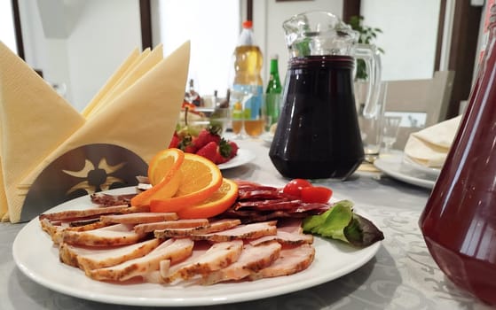 Close view shows meats, oranges, tomatoes, and greens arranged on a plate. Table also includes strawberries, drinks, and napkins. Prepared and ready to serve inside a Restaurant.