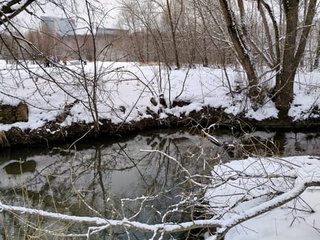 A river flows through a snow-covered park during winter. Bare trees line the riverbank, their branches reflected in the water. Distant figures walk through the snowy landscape in the background.
