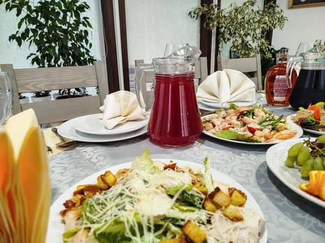 A table is set with plates of food, including salad and fruit, with drinks in glass pitchers. Cloth napkins are neatly folded. Plants provide a background in the Restaurant.