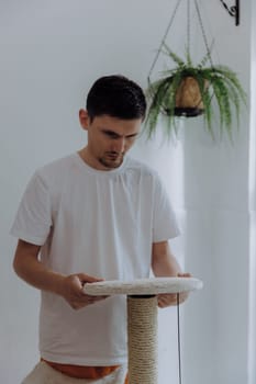 One young handsome Caucasian handsome guy with a serious emotion screws by hand a round shelf to a cardboard tube with a new jute thread to the upper tier of a cat house, after renovation, standing in the kitchen on a summer day, side view close-up with depth of field. The concept of renovating a cat house at home.