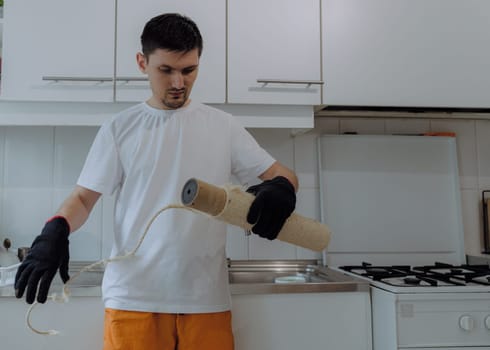 One young handsome Caucasian brunette guy in black gloves unwinds old jute thread from a cardboard tube of a cat house to repair it, stands in the kitchen near the kitchen cabinets on a summer day, close-up side view with depth of field. The concept of renovating a cat house at home.