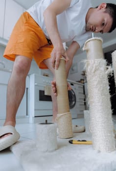 One young handsome brunette guy unscrews a cardboard tube of a cat house with his hands to repair it, standing in the kitchen on a summer day, bottom side view up close up with depth of field. The concept of renovating a cat house at home.