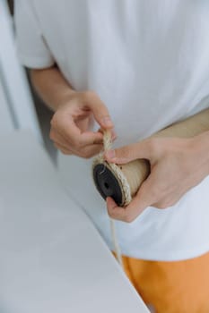 One young Caucasian guy is gluing a new jute thread with hot glue onto a cardboard tube of a cat house, holding it with his fingers and repairing it, standing in the kitchen on a summer day, top side view close-up with depth of field. Concept of repairing a cat house at home.