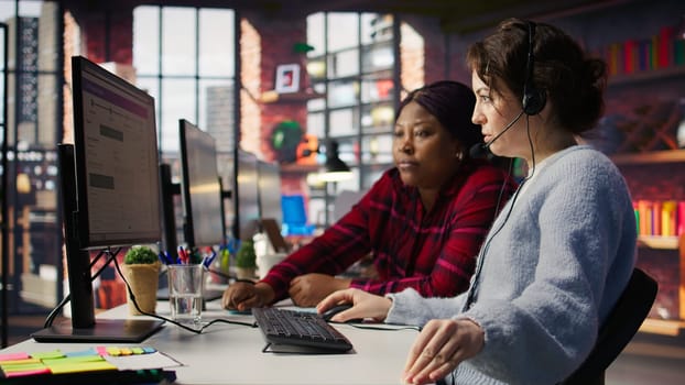 Call center team leader going over paperwork files next to colleague at PC desk. Customer support manager explaining company documentation to intern accommodating with job, camera B