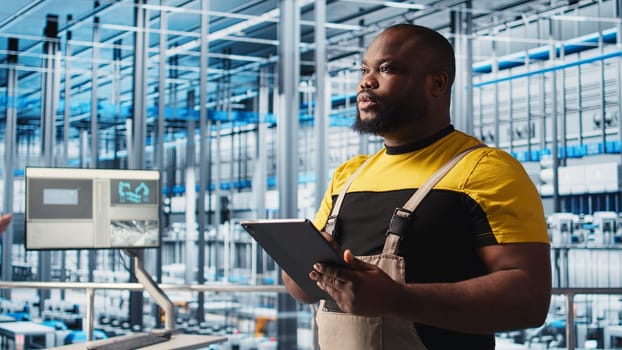 Smart factory engineer oversees operation of robotic arms and assembly lines on facility floor. Industry 4.0 plant african american man monitors system performance and troubleshoots issues, camera A