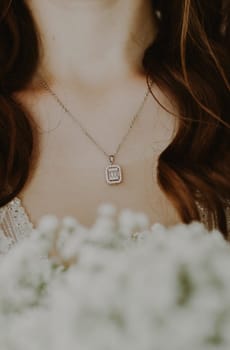 A beautiful chain with a precious pendant hangs on the neck of a young unrecognizable Caucasian brunette bride with a bouquet of white blurry boutonnieres standing on a summer morning in the room before the wedding ceremony, close-up side view.