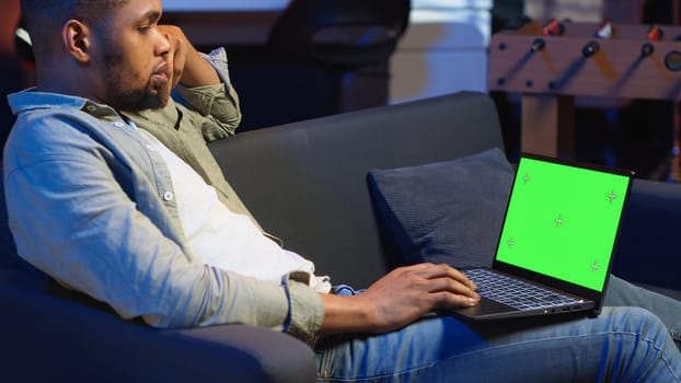 African american young guy using device with green screen from his sofa, enjoys the comfort of his modern cozy home. Casual relaxed person working in the living room, multitasking. Camera B.