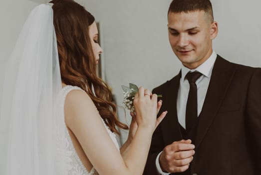 Portrait of one young beautiful stylish white brunette bride inserting a mini bouquet of boutonnieres into the chest pocket of the groom in a dark suit on a summer morning in the room, side view close up. Wedding traditions concept.