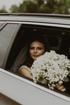Portrait of one young beautiful stylish Caucasian brunette bride with a gentle look sits in a car and looks at the camera and holds out a bouquet of white boutonnieres through an open window on a rainy cloudy day, close-up side view.