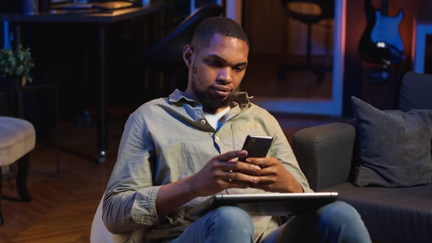 Black man taking a break from work by scrolling his smartphone at home, messaging friends on social media and having fun. Guy relaxing in stylish living room with neon lights. Camera B.