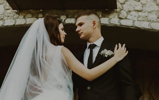 Young handsome Caucasian brunette couple of groom and bride looking at each other with love, holding each other by the shoulders and waist, standing on the steps at the entrance to the wedding castle on a cloudy rainy day, bottom side view close-up with selective focus.