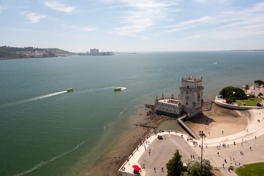 Aerial drone view of Belem Tower on Tagus River with boats creating wakes in the water, highlighting Lisbon's scenic waterfront and historic architecture